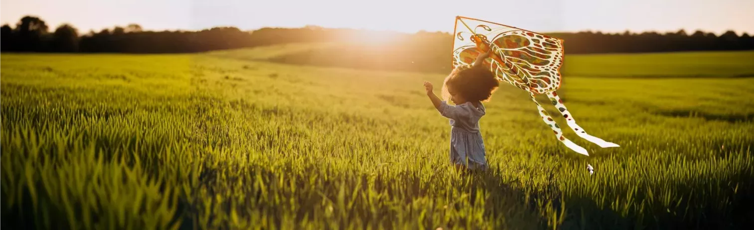 Niña volando una cometa en un luminoso prado verde con el sol de fondo. 